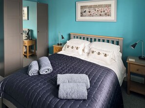 A neatly made bed with a dark blue quilt and pillows, a bedside table with a lamp, a framed picture on the wall, and a door with a mirror.