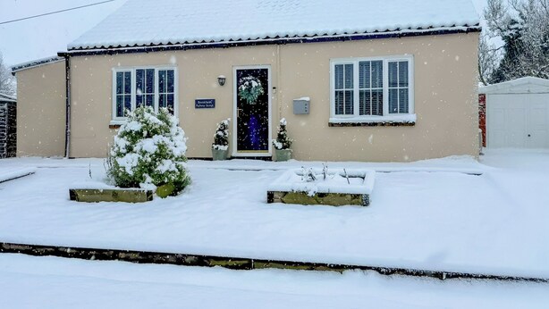 Ein zweistöckiges Gebäude mit schneebedecktem Dach und einer beige farbenen Fassade. Im Erdgeschoss befinden sich zwei Fenster und eine Tür mit einem Schild darüber.