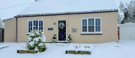 A two-story building with a snow-covered roof and a beige facade. There are two windows on the ground floor and a door with a sign above it.