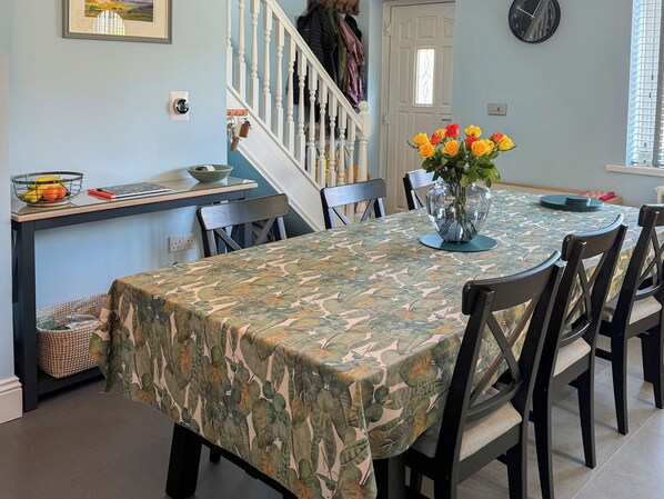 Un coin repas avec une table recouverte d’une nappe à motifs camouflage, entourée de chaises. Un vase rempli de fleurs jaunes est posé sur la table, et un saladier de fruits se trouve sur la table d’appoint.