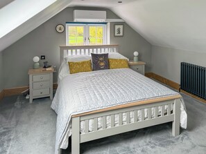 Un dormitorio con techo inclinado, una cama con edredón blanco acolchado, dos mesitas de noche con lámparas, un rabillo y un radiador.