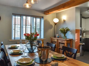 A dining area with a wooden table set for four, a vase with flowers, and a view of the outside through large windows.
