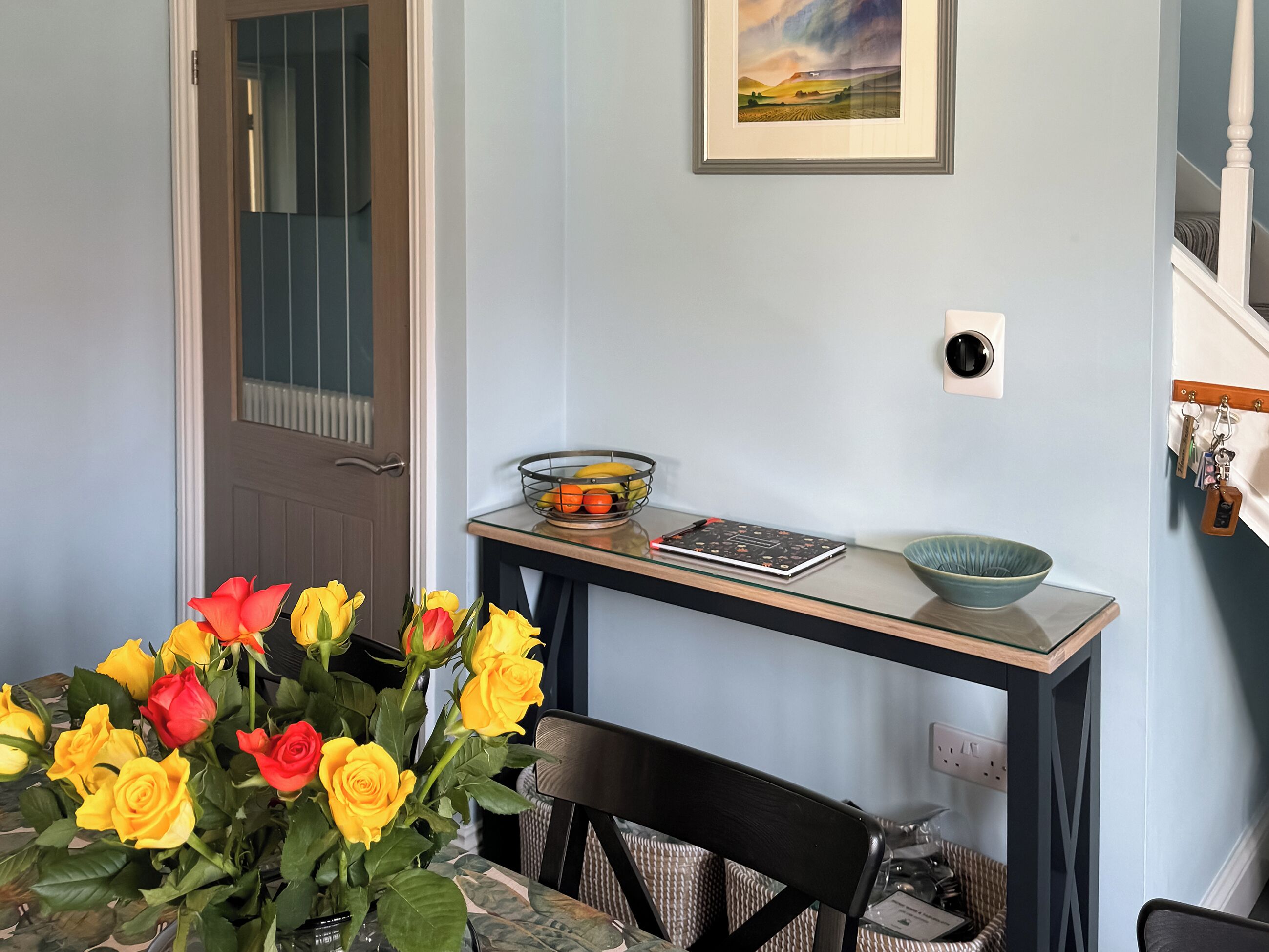 A dining area with a table set for two, a vase of yellow and red roses, a framed landscape painting, a bowl of fruit, and a notebook on a small table.
