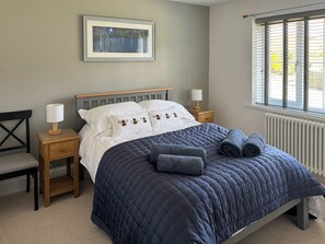 Une chambre à coucher comprenant un lit, deux tables de chevet, une chaise, une fenêtre avec des stores et un tableau encadré accroché au mur.