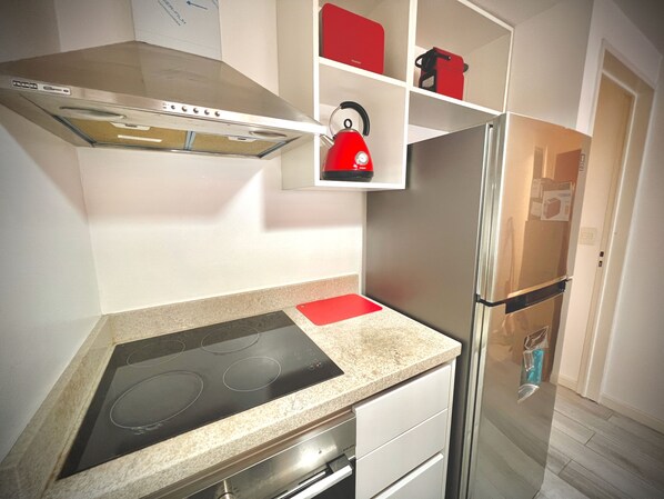 Una cocina moderna con un refrigerador de acero inoxidable, un horno empotrado y una tetera roja sobre una estantería.