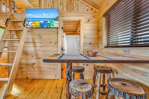 L’intérieur d’un chalet en bois, avec un escalier en bois, un coin repas comprenant une table et des tabourets, et une fenêtre équipée de stores.