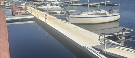 A marina with multiple boats docked, a wooden walkway leading to the dock, and a clear blue sky.