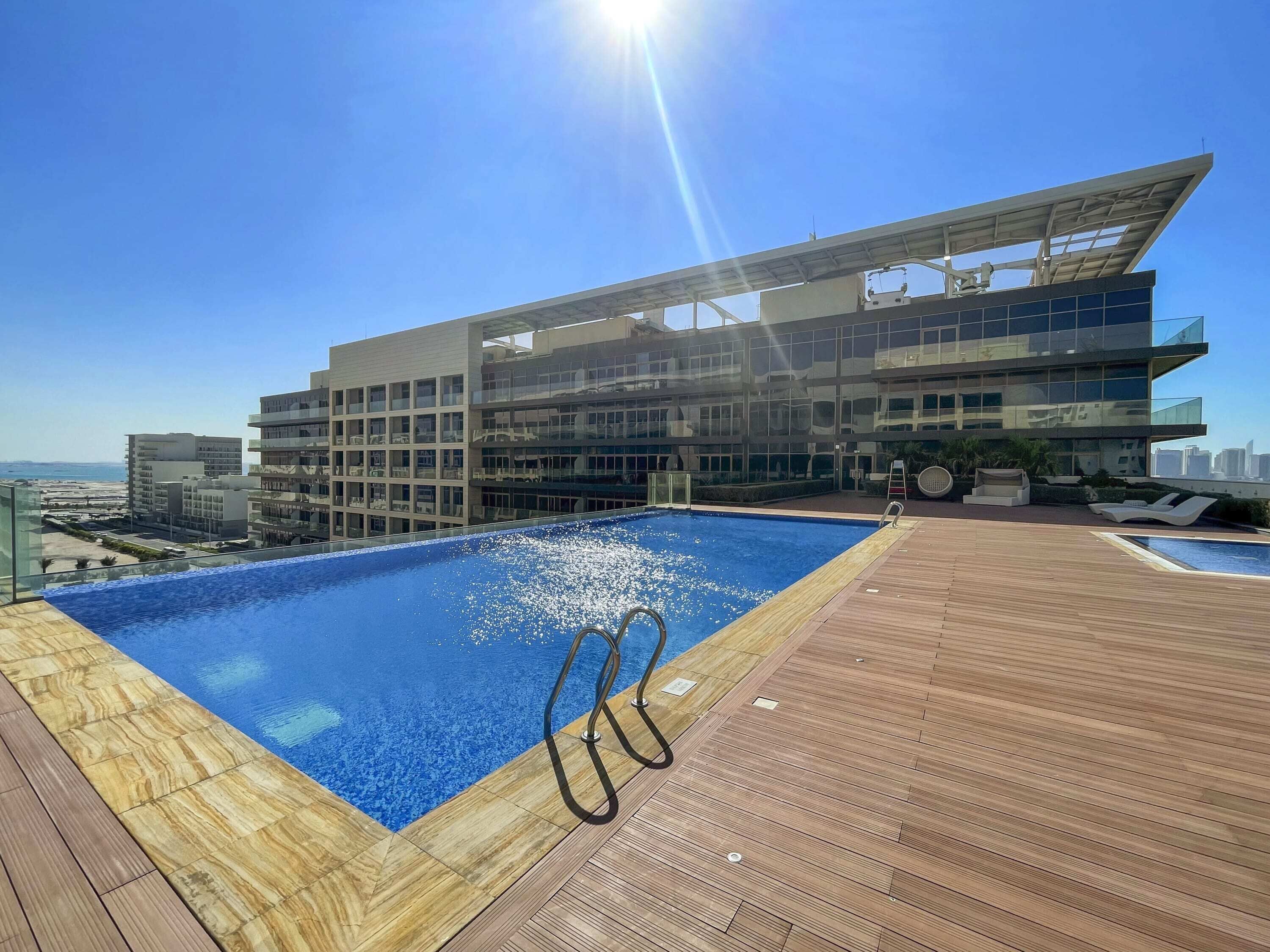 A rooftop pool with a clear view of the city skyline, a modern building with glass balconies, and a wooden deck area.