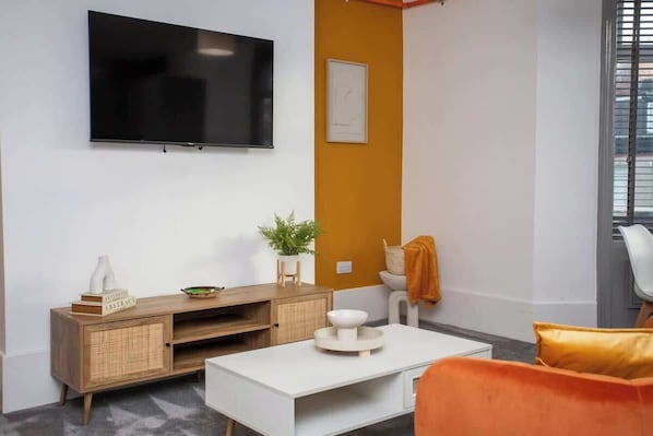 A modern living room with a flat-screen TV, a white coffee table, a wooden cabinet, and a bright orange accent wall.