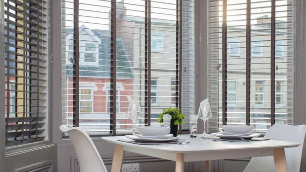 A dining area with a white table set for four, surrounded by white chairs, with a view of buildings through large windows with blinds.