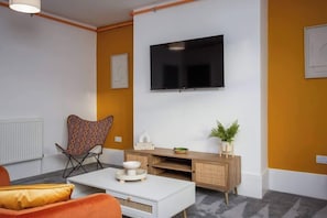 Una sala de estar moderna con un televisor de pantalla plana montado en la pared, una mesa de centro blanca, un sillón estampado, un armario de madera y una planta en maceta.