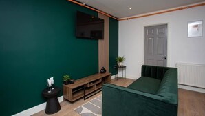 Una sala de estar moderna con una pared verde, un televisor de pantalla plana, un mueble de entretenimiento de madera, un sofá verde, una mesita auxiliar pequeña y una planta en maceta.