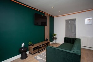 A modern living room with a green wall, a flat-screen TV, a wooden entertainment unit, a green sofa, a small side table, and a potted plant. - Vacant Nests - Atlantic City - Prime Location (Tyne and Wear)