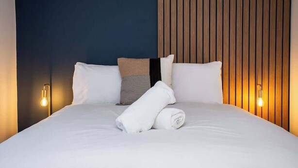 Una cama bien hecha con sábanas blancas y una toalla enrollada, un cabecero de listones de madera y dos lámparas fijadas a la pared.