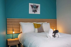 A hotel room with a teal wall, a wooden headboard, a bed with white bedding, a nightstand with a lamp, and a hairdryer on the bed. - Bondi beach Getaway - One bedroom apartment (Tyne and Wear)