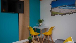 Una stanza con pareti color acquamarina, un pannello in legno, una TV a schermo piatto, un tavolino rotondo con due sedie gialle, una piccola pianta in vaso e un quadro raffigurante una spiaggia.