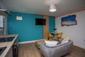 A compact living space with a kitchenette, a grey sofa, a small dining table with yellow chairs, a mounted TV, and a wall-mounted air conditioner. - Bondi beach Getaway - One bedroom apartment (Tyne and Wear)
