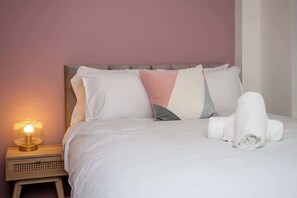 Ein ordentlich bezogenes Bett mit Kissen, einer Nachttischlampe und einem aufgerollten Handtuch.