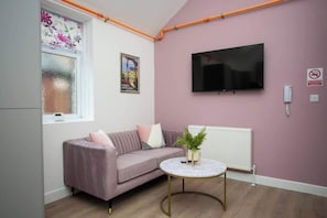 Une pièce avec un mur rose, un téléviseur à écran plat fixé au-dessus d’un radiateur blanc, un canapé avec des coussins, une petite table ronde avec une plante, et une fenêtre avec du verre dépoli et des motifs floraux.