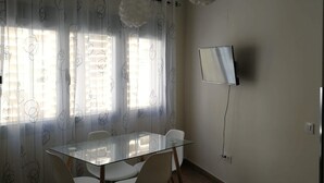 Un comedor con mesa y sillas de vidrio, televisor montado en la pared y una ventana con cortinas traslúcidas.