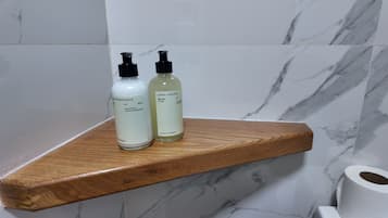 A wooden shelf with two bottles of liquid soap and a roll of toilet paper against a marble wall.