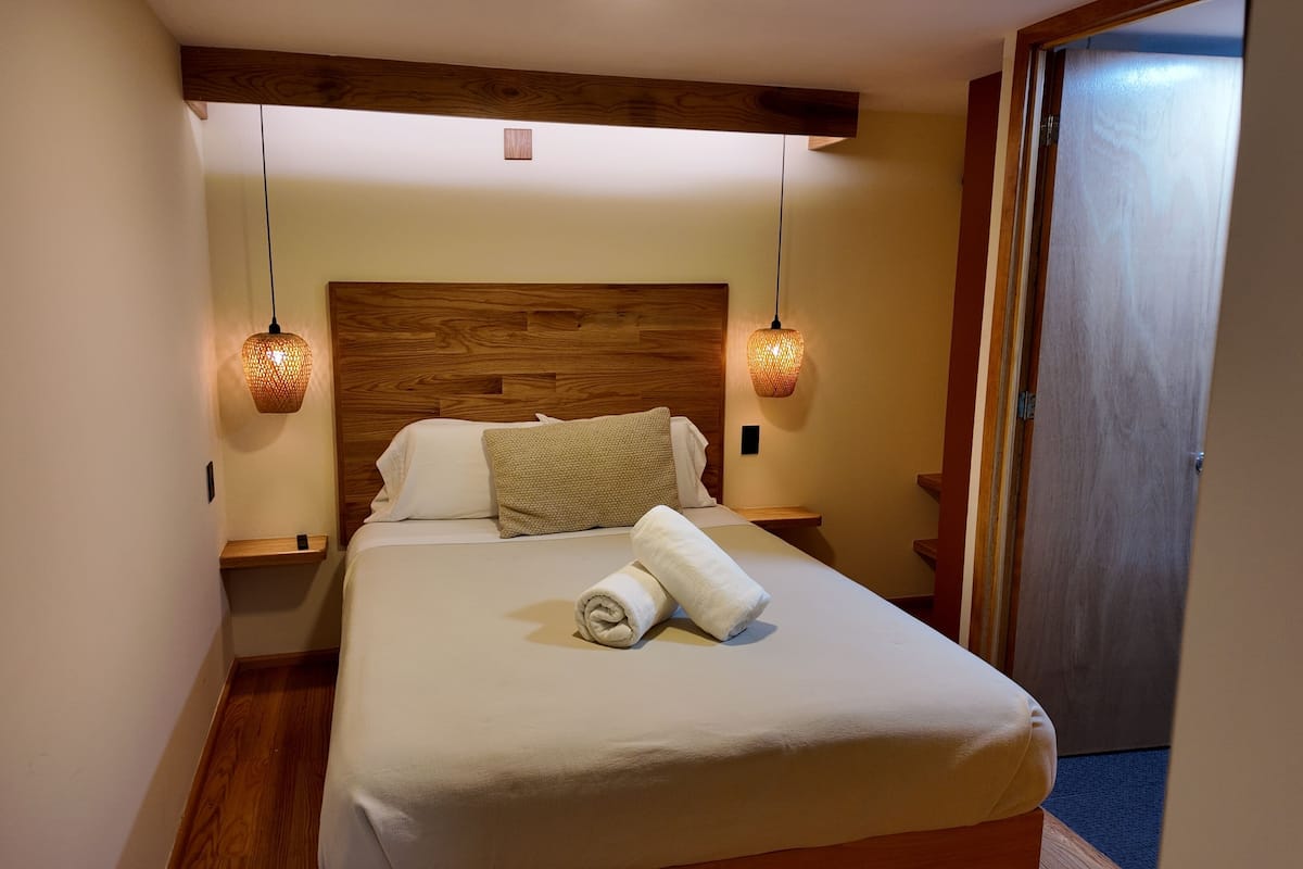 A neatly made bed with a rolled towel, wooden headboard, and two wall-mounted lamps.