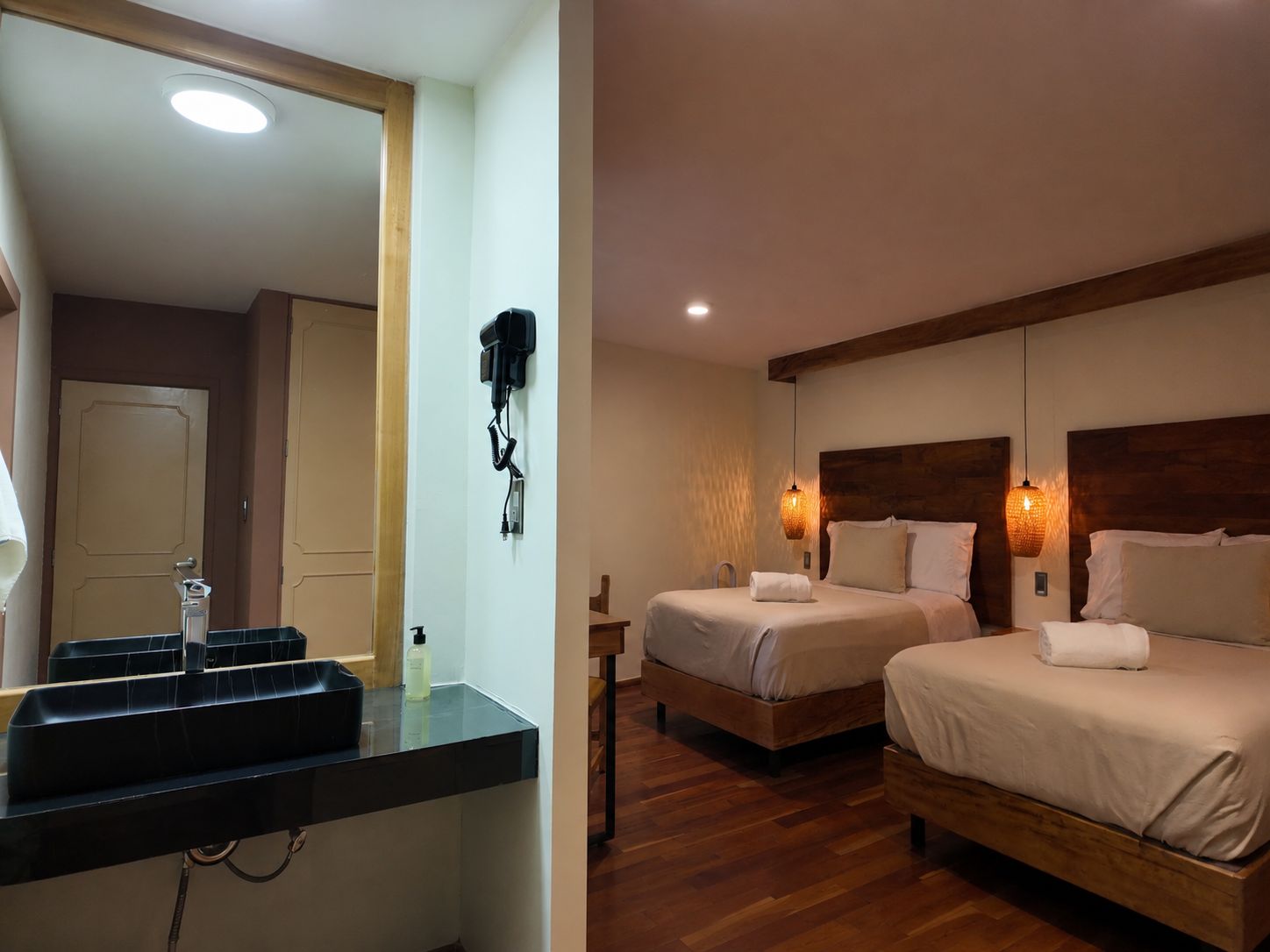 A hotel room with a wooden floor, two beds with white linens, a bathroom with a black countertop, and a mirror.