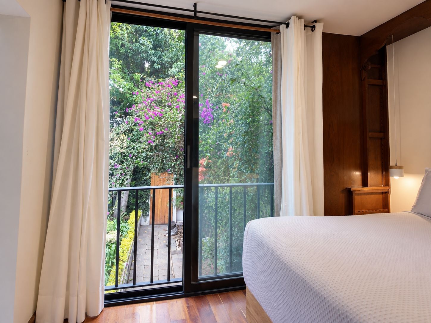 Un dormitorio con un amplio ventanal corredera que da a un balcón con vistas a la vegetación y flores.