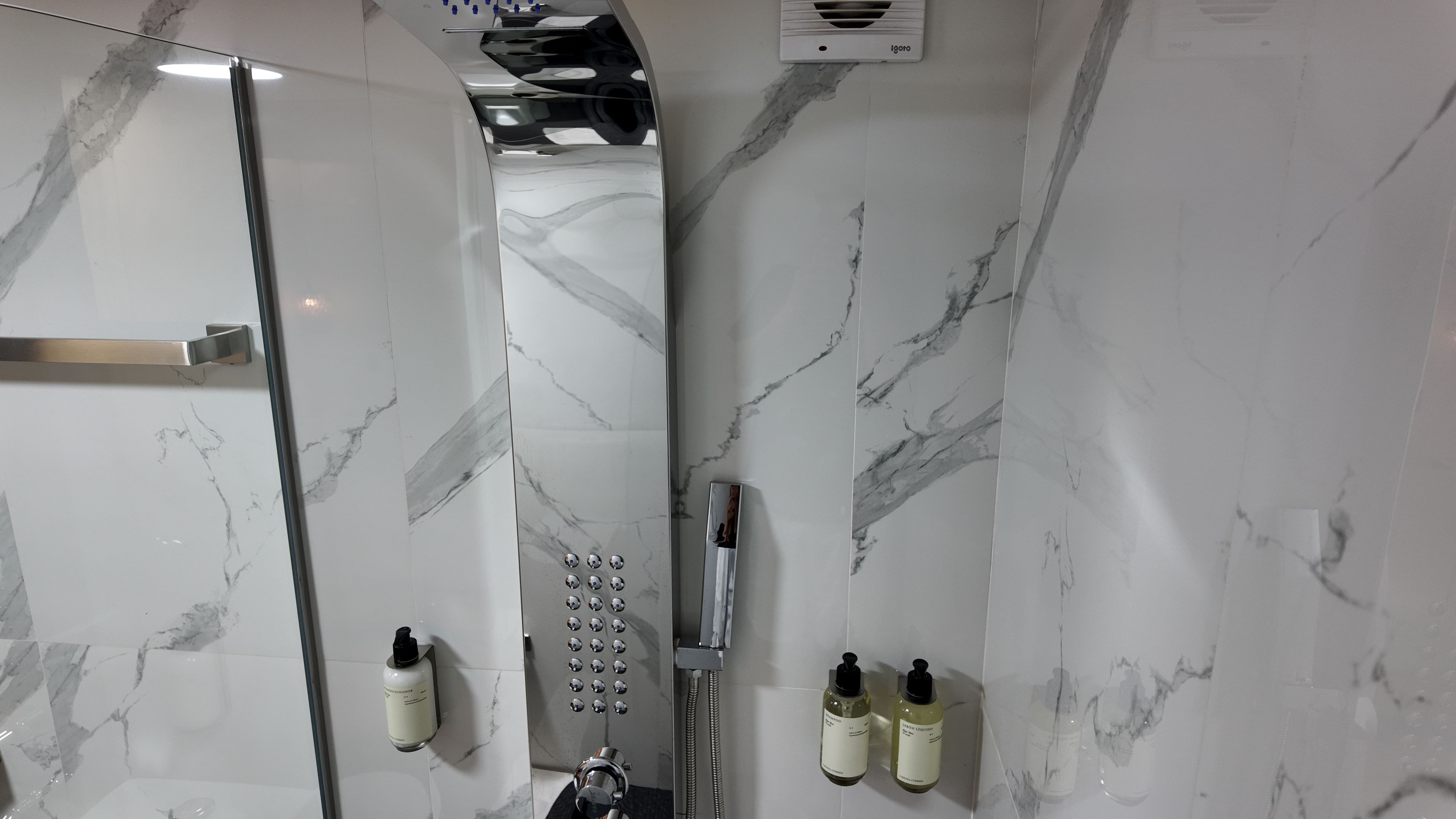 A modern bathroom with a glass-enclosed shower, marble walls, and toiletries mounted on the wall.