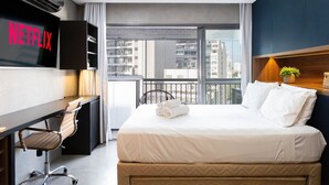 A hotel room with a bed, a desk with a chair, a television displaying 'NETFLIX', and a view of the city through large windows. - Condomínio Setin Downtown Genebra (São Paulo)