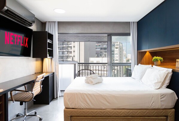 Une chambre d’hôtel avec un lit, un bureau et une chaise, un téléviseur diffusant du contenu « NETFLIX », et une vue sur la ville grâce à de grandes fenêtres.
