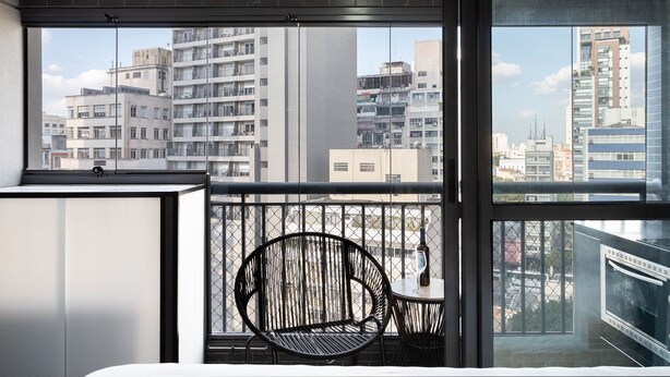 A modern room with a large window, a black chair, and a small table with a bottle. - Condomínio Setin Downtown Genebra (São Paulo)