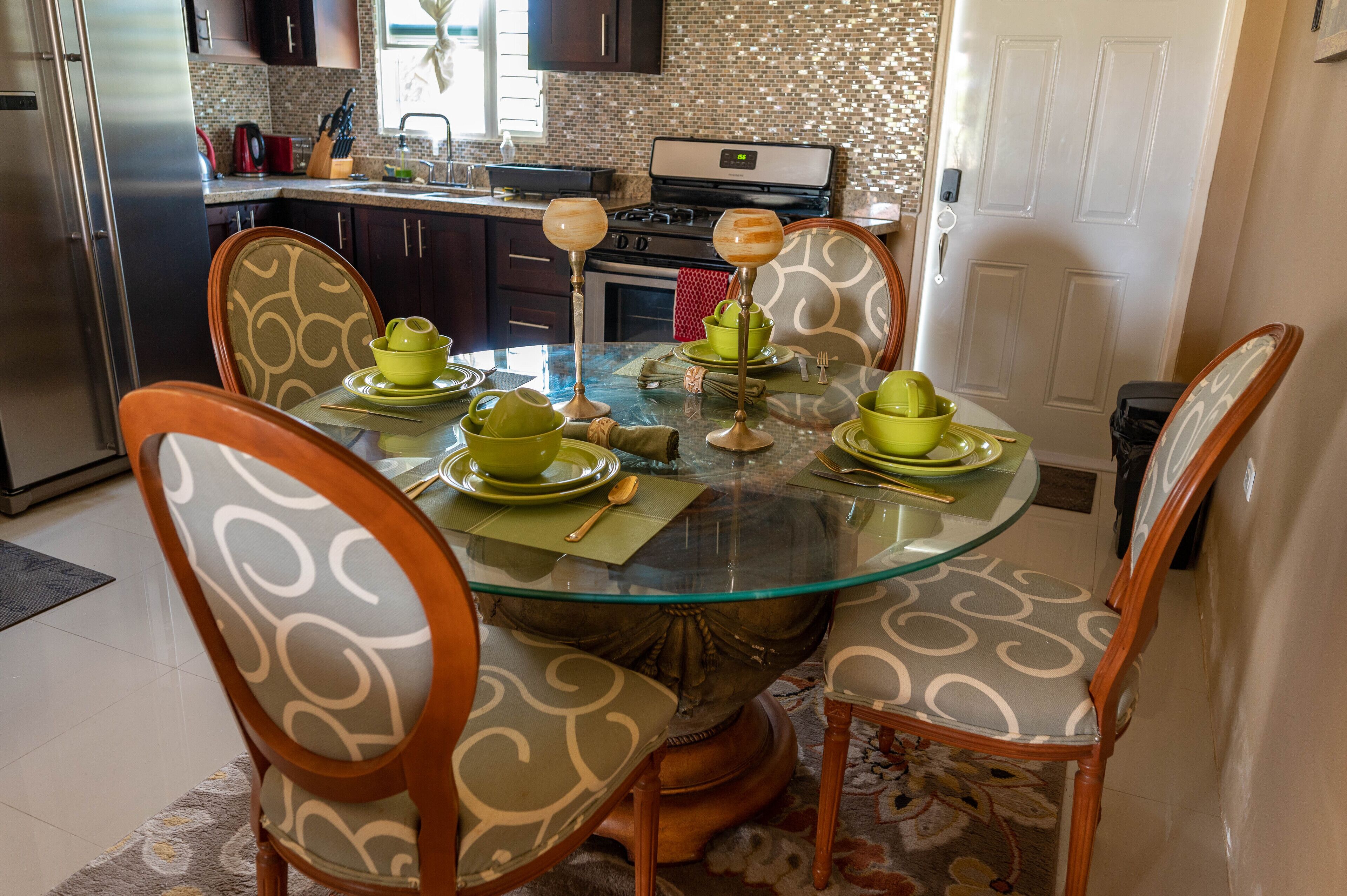 Uma área de refeições com mesa de vidro rodeada por cadeiras estampadas, cozinha ao fundo e painel de azulejos.