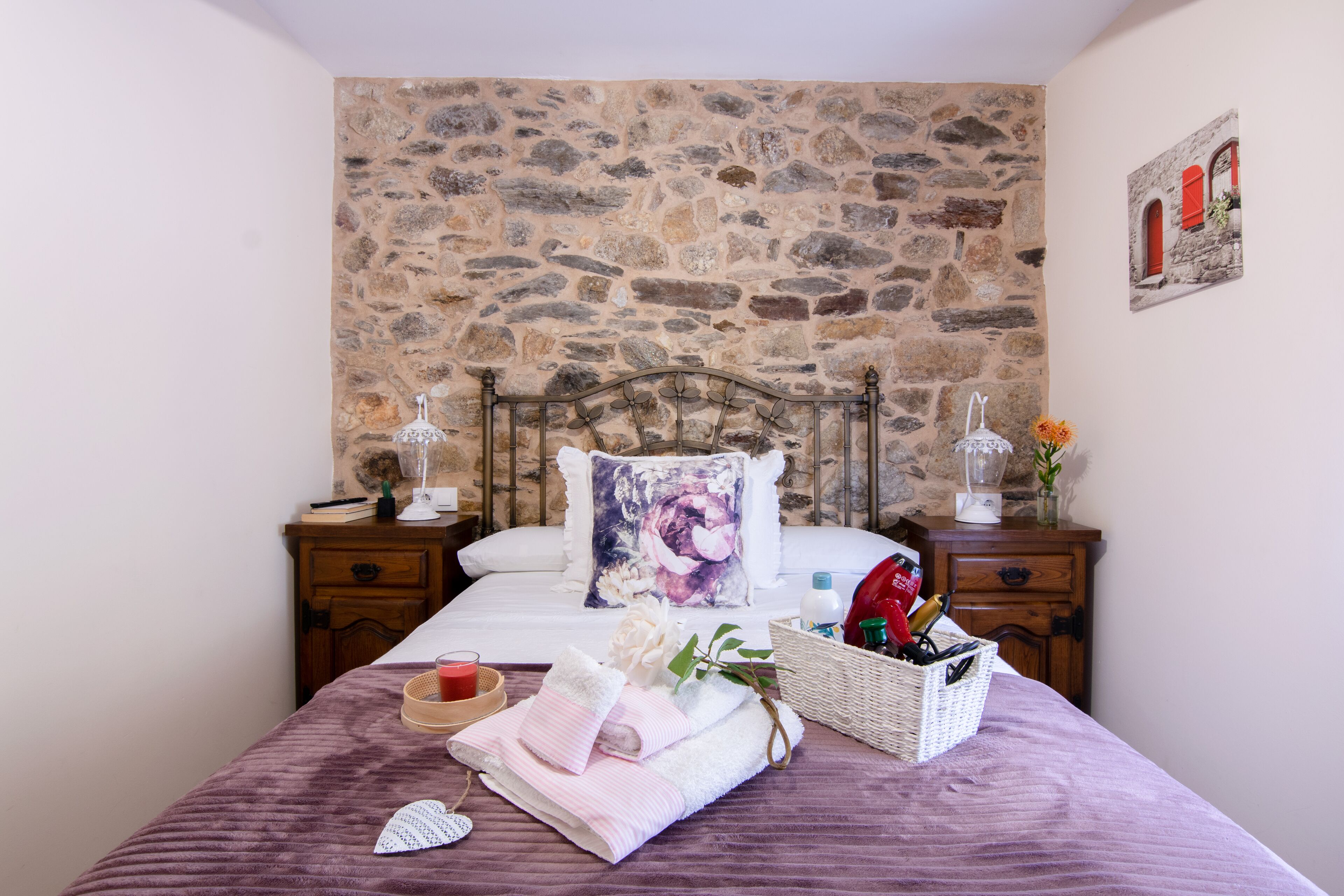 Un dormitorio con pared de piedra, una cama con almohada estampada, mesitas de noche con lámparas, un cesto de artículos de aseo y una planta pequeña.
