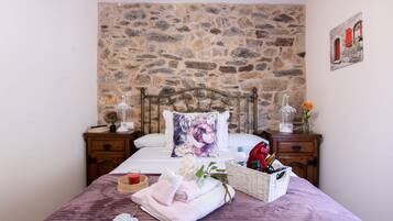 Un dormitorio con pared de piedra, una cama con almohada estampada, mesitas de noche con lámparas, un cesto de artículos de aseo y una planta pequeña.