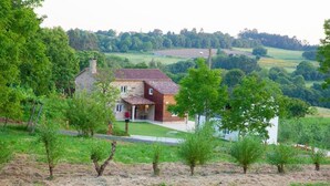 Una casa rural con fachada de piedra y un ala de techo rojo, rodeada de vegetación y campos.