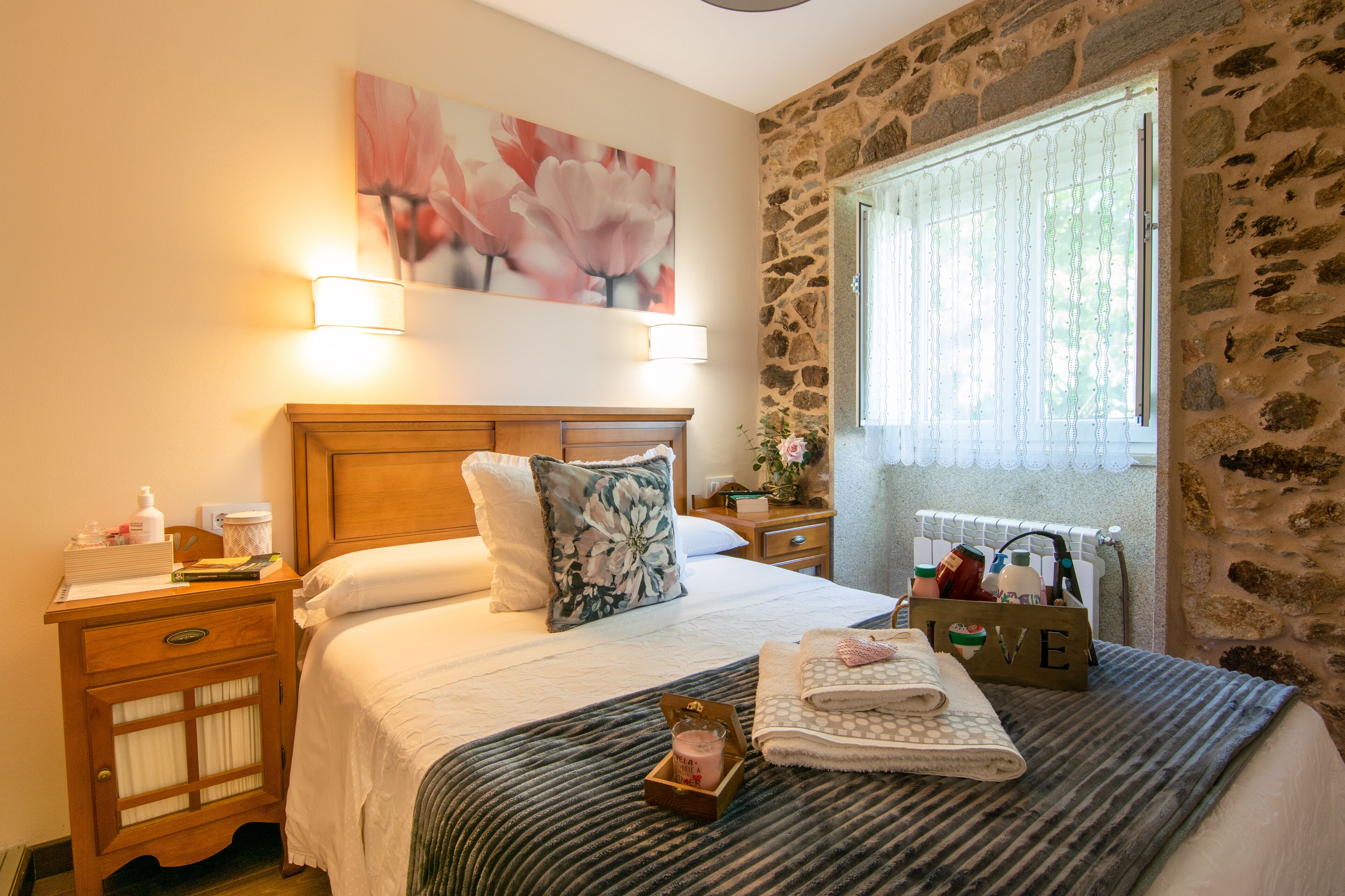A neatly made bed with a floral pillow, a bedside table with a lamp, a nightstand with a lamp, and a window with a view of greenery.