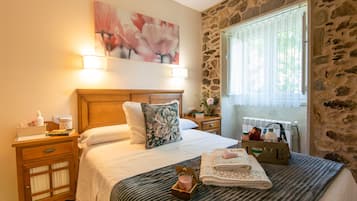 A neatly made bed with a floral pillow, a bedside table with a lamp, a nightstand with a lamp, and a window with a view of greenery.