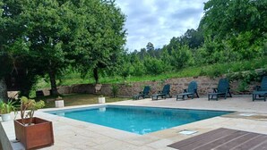 Una piscina rectangular con solárium de piedra, rodeada de árboles verdes y un muro de piedra.