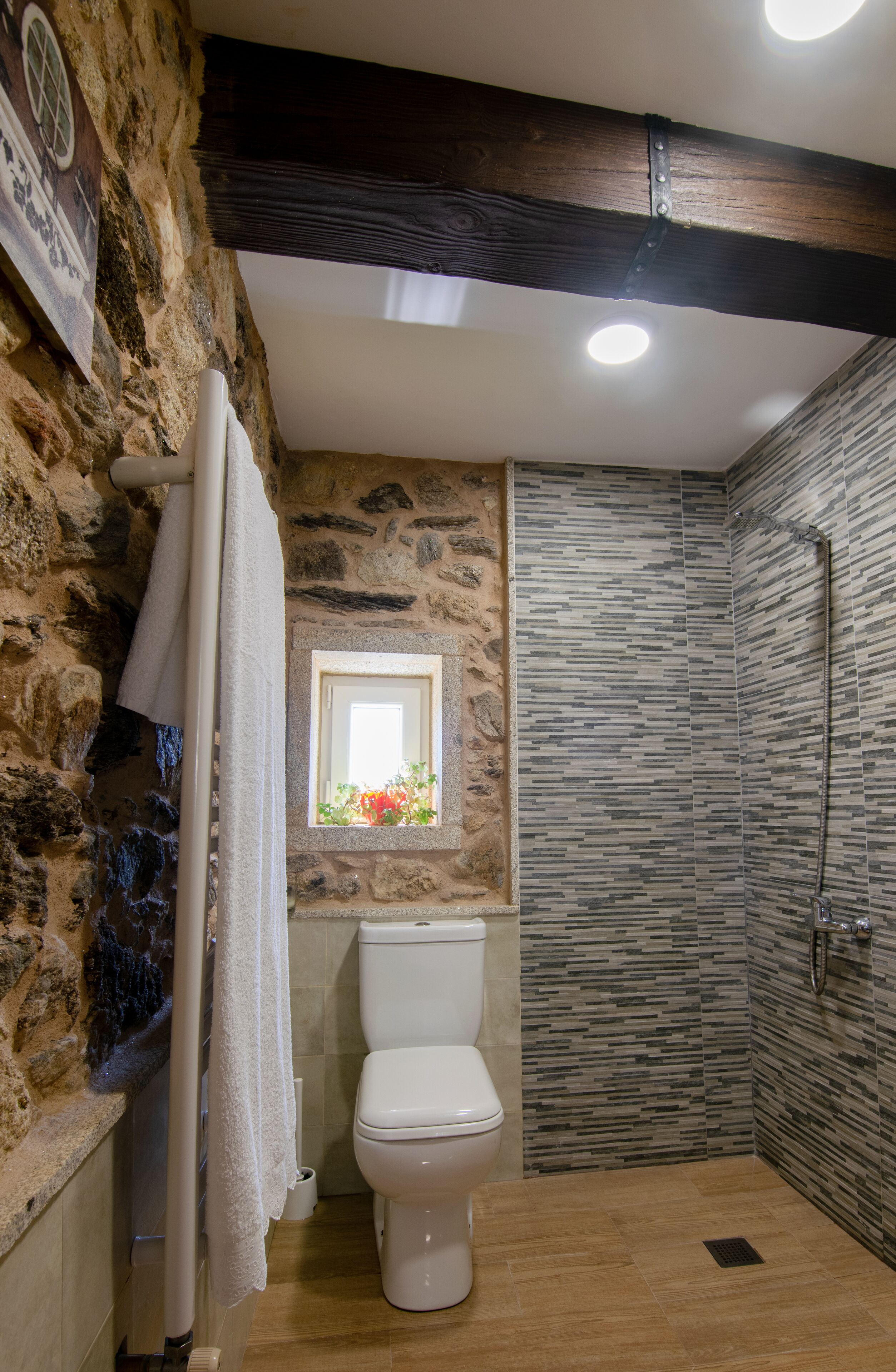 Un baño con pared de piedra, techo de vigas de madera, inodoro blanco y zona de ducha.