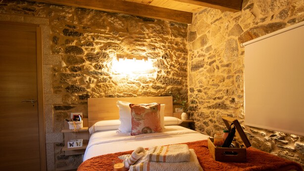 Habitación de hotel con pared de piedra, techo de madera, cama con manta roja, mesita de noche con una vela y una caja de madera con artículos de aseo.