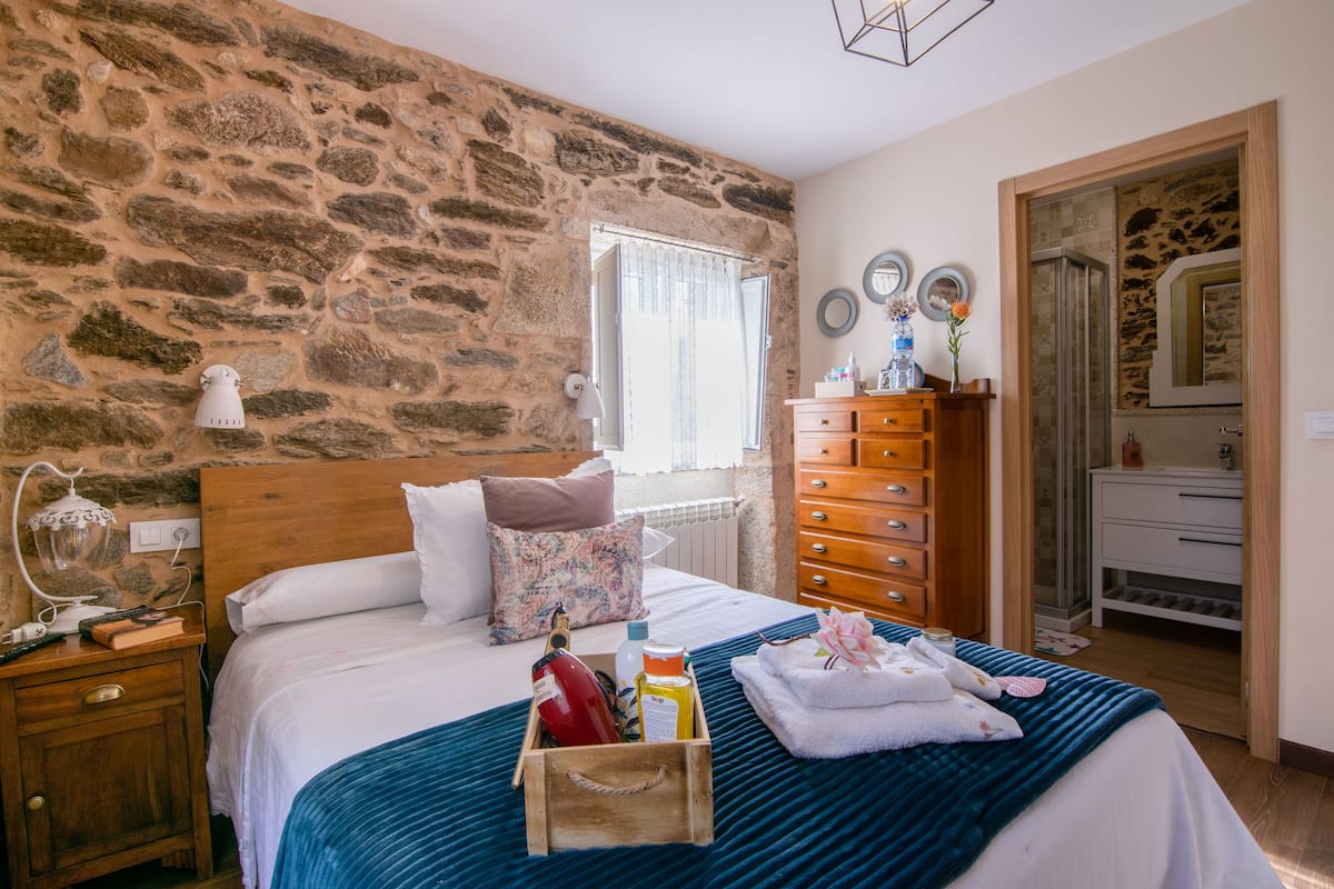 A bedroom with a stone wall, a wooden headboard, a bed with a blue bedspread, a wooden dresser, and a nightstand with a lamp.