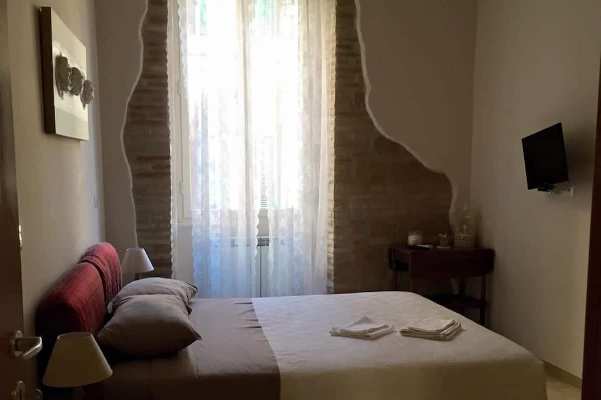 Een slaapkamer met een groot bed, een nachtkastje met een lamp, een klein tafeltje met een vaas, en een televisie aan de muur.