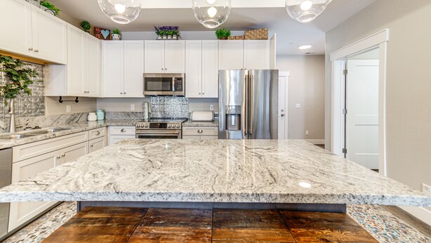 A modern kitchen with white cabinets, a large granite island, stainless steel appliances, and pendant lighting.