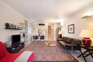 A living room with a sofa, TV, and a coffee table. There is a dining area with chairs and a table. The room has a patterned rug and a lamp on a side table.
