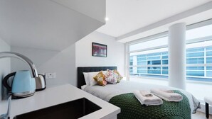 A modern hotel room with a large window, a bed with a green blanket, a kettle, and a painting on the wall.