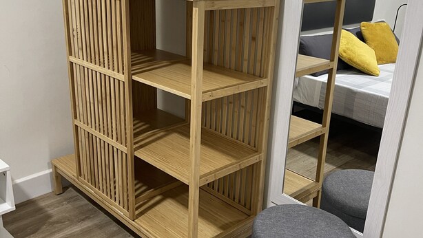Une pièce avec une armoire en bois, un pouf gris et un lit avec des coussins jaunes.