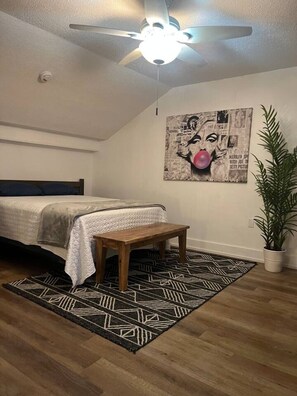 Una camera da letto con un letto, una panca di legno, una ventola a soffitto, una lampada a parete, un tappeto decorato e una pianta in vaso.