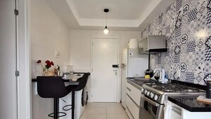 Eine kompakte Küche mit Gasherd, Backofen und Mikrowelle. Es gibt einen Essbereich mit Tisch und zwei Stühlen. Die Küche hat eine gemusterte Spritzschutzwand und eine Deckenleuchte.