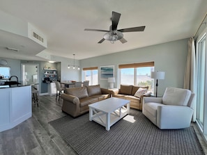 A modern living room with a sofa, armchair, and coffee table, a dining area with chairs, and a kitchen with a bar. - CALYPSO BEACHFRONT 3BR/3BA 3rd Flr CRNR! 4 BEACH CHAIRS INCLUDED! See Reviews! (Panama City Beach)